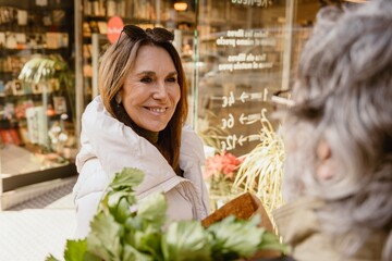 A White elder woman with sunglasses on her forehead wearing a white winter coat and smiling warmly while talking to her husband outside a bookstore, holding leafy greens and a paper bag.