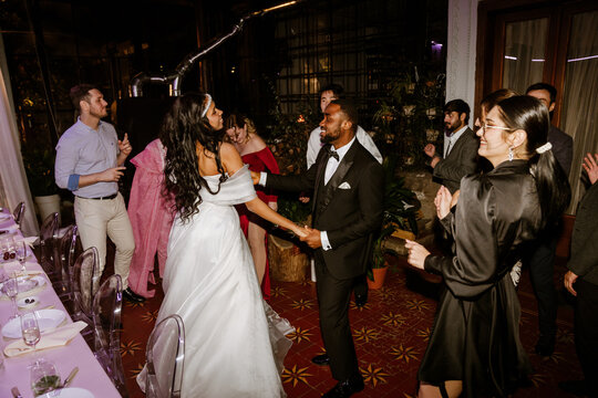 A Black young bride and groom dancing together on a red tiled floor surrounded by a multinational group of wedding guests celebrating during a lively indoor wedding reception with plants.