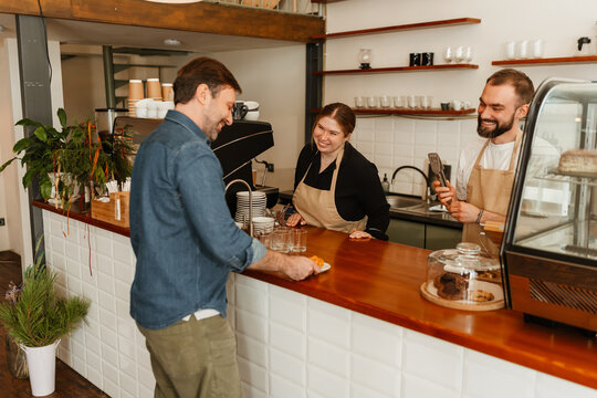 A white adult man receives food at a coffee shop counter from two smiling cafe workers, a woman and a man wearing aprons. They are engaged in a friendly conversation.