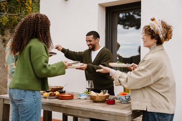 Three adults stand at a table on the veranda, filled with food. In the center, a smiling Black man...
