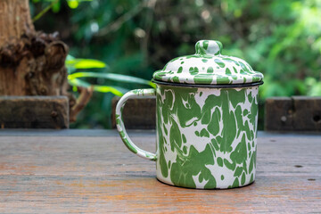 Green and white enamel mug or cangkir blirik with a lid, resting on a wooden table outdoors, with...