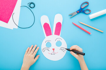 Little child hand drawing rabbit paper mask with marker on light blue table background. Pastel...