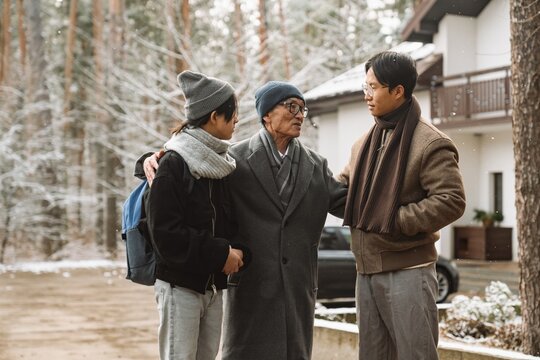 In the winter yard near the house an Asian father of about 60 years old stands between his teenage son and son of about 20 years old and hugs them. Concept of fatherhood, talking with children.