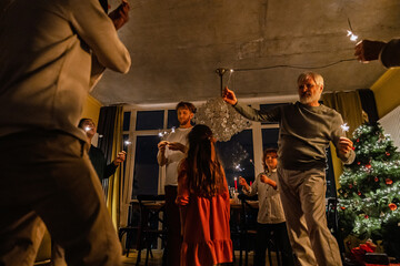 A White father, mother, and two children hold lit sparklers while dancing near a Christmas tree in a room with white windows and, a concrete ceiling. The grandfather with white hair is in the middle.