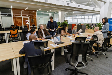 A European businessman in his 30s, smiling while standing next to a table of his colleagues, talking to his older, mid-50s male colleague, in an office hall