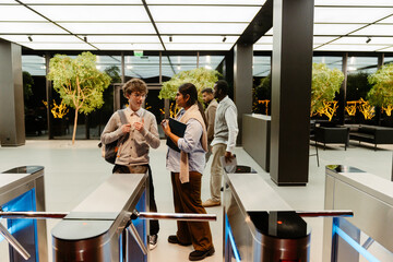 In a spacious modern office lobby near electronic turnstiles, a White man, an intern in his 20s, talks to an Indian woman in her 30s holding a folder, while two other men walk by in the background.