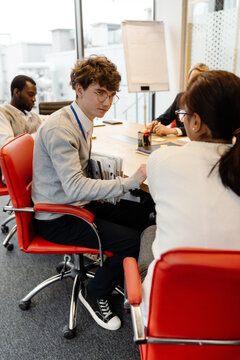 A Caucasian male intern aged 25, sitting at a desk among his colleagues in an office meeting room, and turning to talk to his young female colleague who sits beside him, with windows all around them