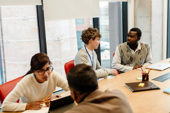 A Black man in a sweater vest and a White man in a gray cardigan in their 20s, talk while sitting at a wooden table near an Indian woman and a man talking, in an office meeting room with big windows.