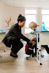 Fototapeta premium A Black female teacher in her late 30s, dressed in a black business suit, helping her young, first-grade White male student pack his backpack as he sits at his desk in a classroom