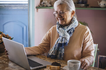 Happy white haired senior woman wearing eyeglasses sitting indoor using laptop while has a coffee break, addicted  elderly lady enjoying food and drink