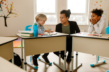 A Black woman teacher in her 30s wearing a black blazer crouches near and looks at a smiling White school-aged boy drawing at his desk, next to a Black school-aged boy who looks at them in a classroom