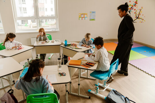 A group of diverse elementary school students sit at desks in a bright classroom drawing and writing, while an adult Latino female teacher observes them. The classroom has white walls and windows.