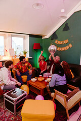 A multiethnic group of friends in their 20s are at a home birthday party, sitting around a table with snacks, two are a White man holding a disco ball, and an Asian woman pointing at the ceiling.