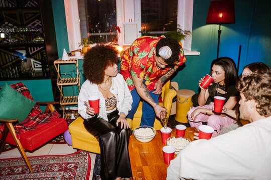 Multiethnic friends in their 20s party in a colorfully decorated living room where a tattooed Latino man wearing a hat pours a drink into red cups on the table with bowls of popcorn where people sit.