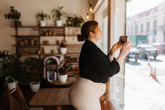 A side view of a smiling brunette White woman with a forearm tattoo wearing a black shirt and white pants in her late 20s opening a cafe by flipping a small wooden sign hanging on the large window. - Powered by Adobe
