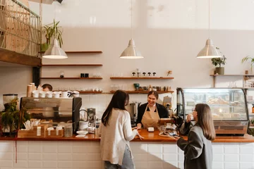 A White, overweight female barista aged 25, taking the order from two women in their late 20s, with a wide smile on her face, as she stands behind the counter in a cozy coffee shop © Drobot Dean