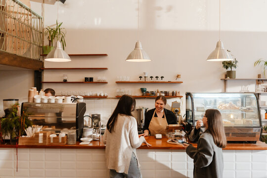 A White, overweight female barista aged 25, taking the order from two women in their late 20s, with a wide smile on her face, as she stands behind the counter in a cozy coffee shop
