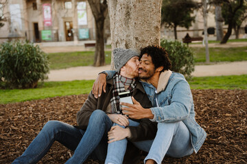 Fototapeta premium An African American man aged 35, wearing a denim jacket, sitting on a field in a park with his mid-30s White husband, and taking a selfie with his phone as the husband kisses him on the cheek