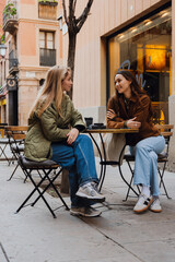 An adult blonde White woman in a green jacket and a brunette White woman in a brown coat sit at a cafe table outside. They converse with a tree and building facade captured in the background.