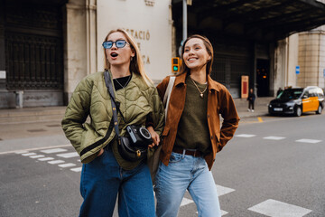 Fototapeta premium Two young European women aged 25, a blond, and a brunette, walking across a road together in the city and looking to the side as the blond has an amazed expression on her face, on a sunny day