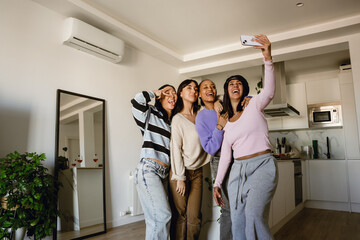 Fototapeta premium A mid-20s Latina woman wearing a stylish black hat, taking a selfie with a group of her three female friends on her phone, as they stand beside her, in her home, near the kitchen counter