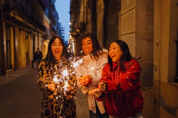 Three young women of different ethnicities stand in a dimly lit alley at night. One wears a leopard print coat, another a white sweater, and the third a red jacket. They hold sparklers and celebrate.