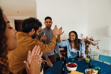 A 25-year-old Black woman with long brunette hair, high-fiving her mid-20s Arabic male friend as they stand among other friends near a table set for a small party, in a white apartment