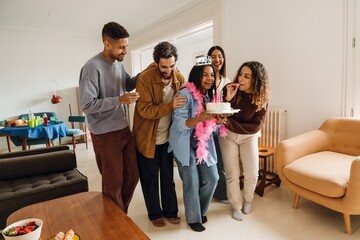 In a sunlit living room, five multiethnic smiling friends in their 20s celebrate the birthday of a Black woman holding a cake wearing a tiara and pink boa, with other people standing around her.