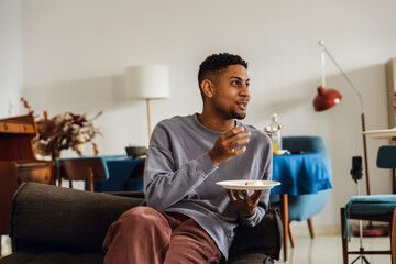 At home, a smiling brunet African American man in his early 30s, wearing a gray sweatshirt and red pants, is sitting on a small black couch, eating a piece of cake, and talking to someone to the side.