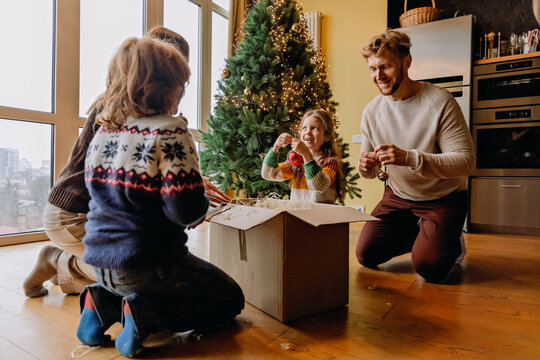 A European family of four, mid-40s parents and their two school-aged kids, a boy, and a girl, unpacking a box of Christmas lights while sitting on the floor of their home, next to the decorated tree