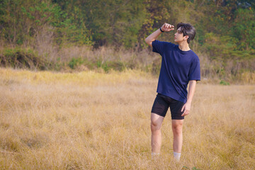 Fototapeta premium Handsome young man in a blue shirt and black sports shorts is happily and relaxing on a morning after-morning exercise, standing and posing on a green lawn in the park. Healthy exercise.
