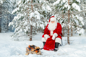 Fototapeta premium Authentic Santa Claus with a bag of gifts sits near a fire in a snowy forest.