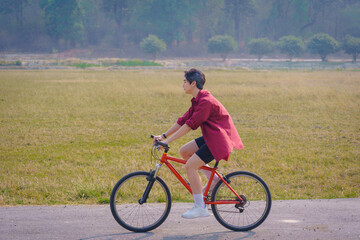A Handsome young man in a red shirt and black sports shorts is happily riding a red bicycle in the morning for exercise and relaxing in the park during holiday. Healthy exercise concept.
