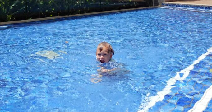 Playful toddler stands in shallow wading pool, taking mouthful of water and puffing up his cheeks. With cheeky grin, he squirts water in our direction, splashing playfully.