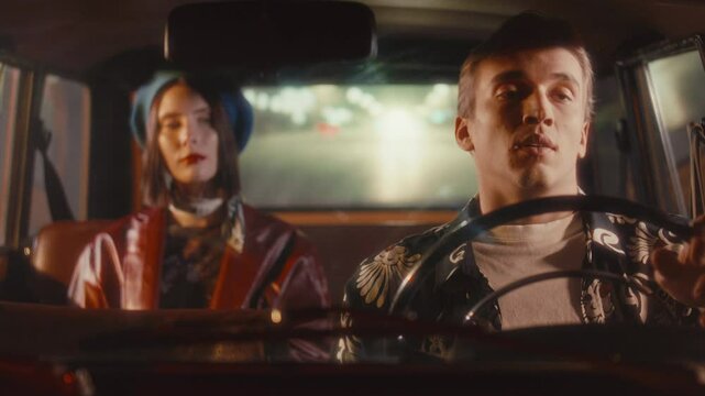 Young man driving vintage car with contemplative female passenger in blue beret and leather jacket sitting in back seat during night ride through city street