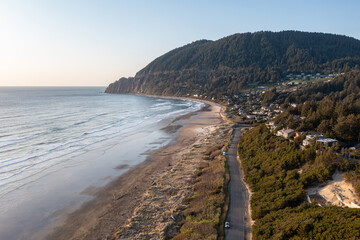 Aerial drone picture of Manzanita Beach on the Oregon Coast, showing waves of the Pacific Ocean, small coastal town, sunset, sandy shoreline, forested mountains. Perfect for travel and tourism themes