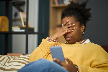 African American woman wearing a yellow sweater sitting on a couch and holding phone with one hand,...