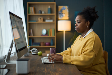 African American woman working at a desk with a desktop computer in a home office setting, shelves with books and decorative items in the background
