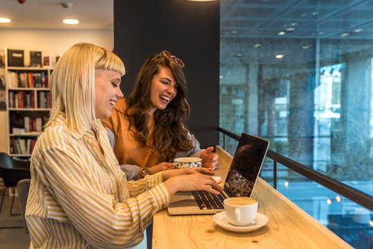 Cheerful young adult student girl helping college friend with research study in library. Two happy trainees working at laptop, watching online presentation on computer, smiling, laughing. Education