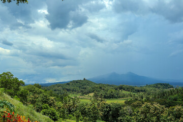 A wide-angle shot of a lush green forest valley stretching to a distant mountain range, with a dramatic sky of dark, stormy clouds overhead.