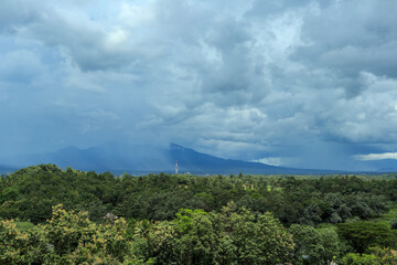 A wide-angle shot of a lush green forest valley stretching to a distant mountain range, with a dramatic sky of dark, stormy clouds overhead.