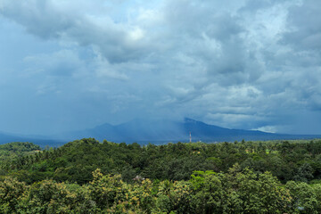 A wide-angle shot of a lush green forest valley stretching to a distant mountain range, with a dramatic sky of dark, stormy clouds overhead.