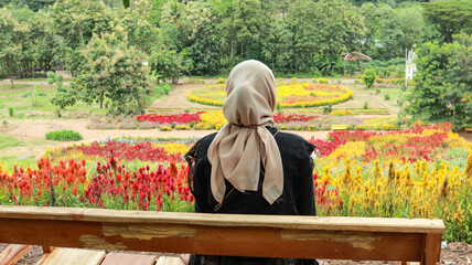 A woman wearing a beige hijab sits on a wooden bench, gazing out at a colorful flower garden.