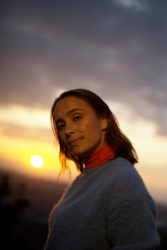 A woman stands against a sunset backdrop in a sweater with the sun setting below clouds.