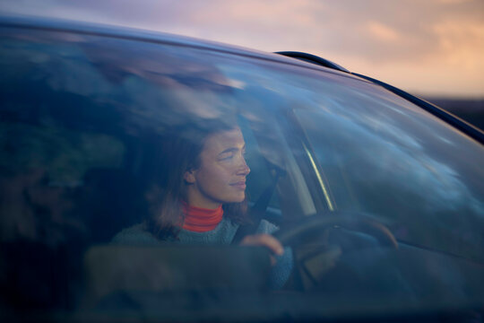 Woman driving a car at sunset with a thoughtful expression, seen through a windshield.