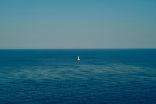 A lone sailboat glides across a calm, vast blue ocean under a clear sky. Peloponnese, Greece