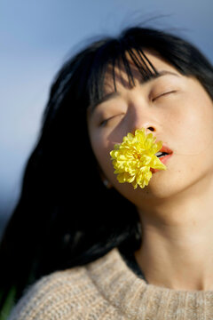 Woman with closed eyes holds a yellow flower in her mouth against a blurred blue background. Brandenburg, Germany
