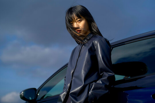Woman in dark coat stands confidently beside a shiny car under a vivid blue sky. Brandenburg, Germany