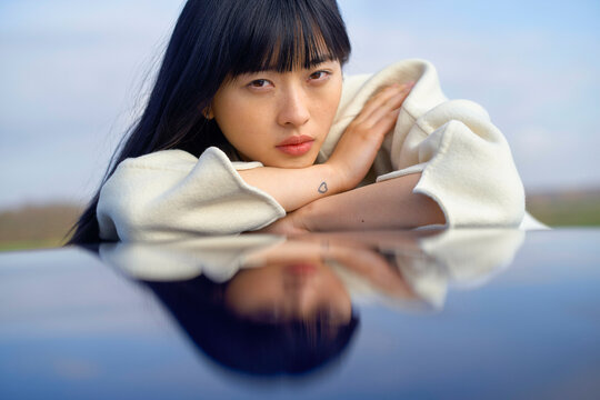 Woman with black hair resting on arms, reflected on a shiny surface with a calm sky backdrop. Brandenburg, Germany