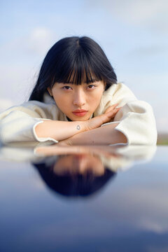 Woman with black hair leaning on a reflective surface, with a distant sky background. Brandenburg, Germany